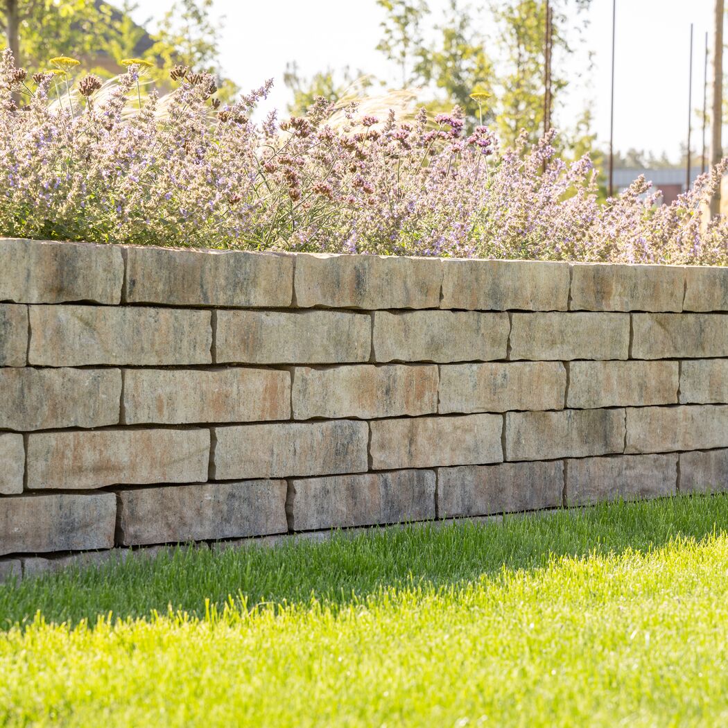 Mauerstein 'T-Wall Castra' muschelkalkfarben mit Blumen und Gras.