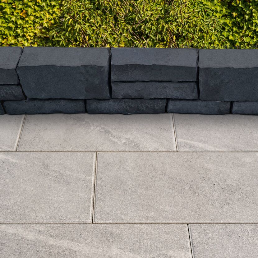 Basaltfarbener Mauerstein 'T-Wall Castra' mit heller Pflasterung und Hecke.