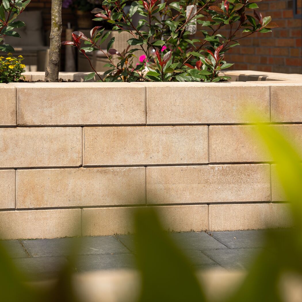 Sandsteinfarbene Mauersteine 'T-Wall Fine Maxi' mit Pflanzen im Hintergrund.