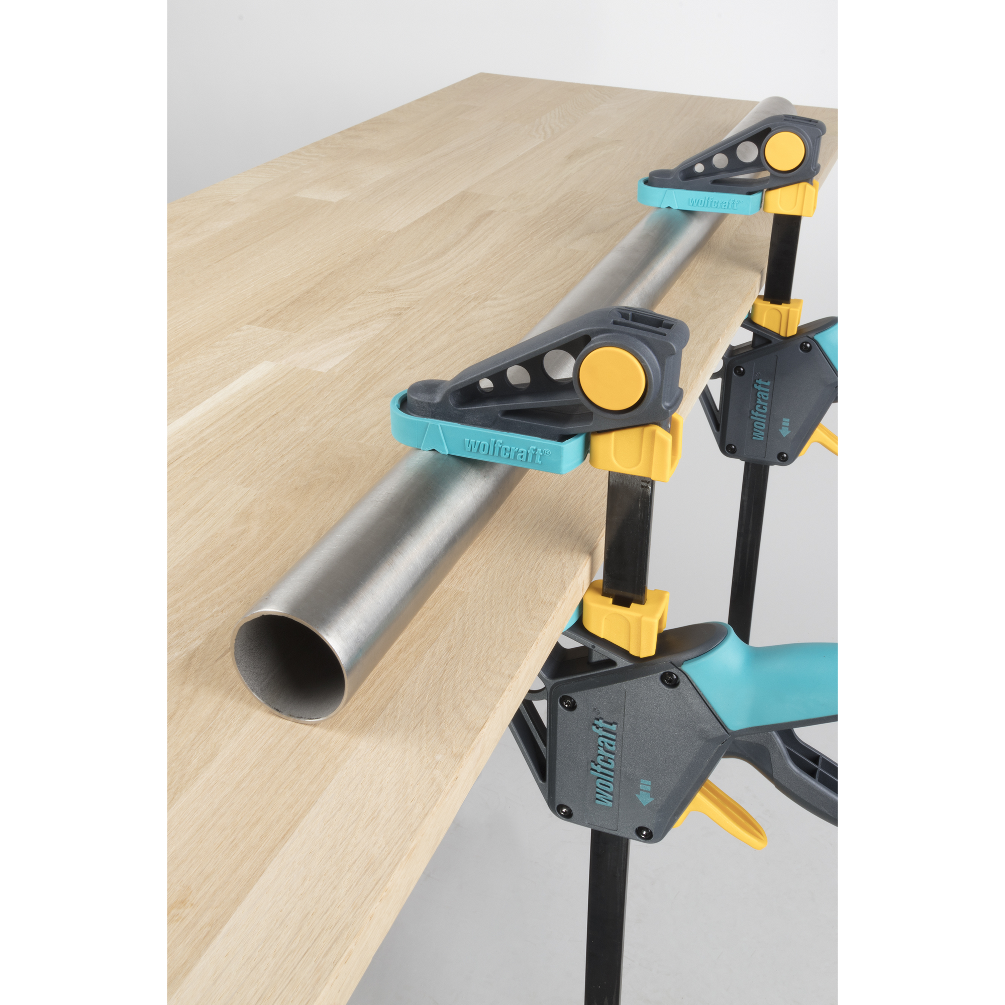 Zwei Einhandzwingen Wolfcraft Proaktiv fixieren ein Metallrohr auf einem Holzbrett.