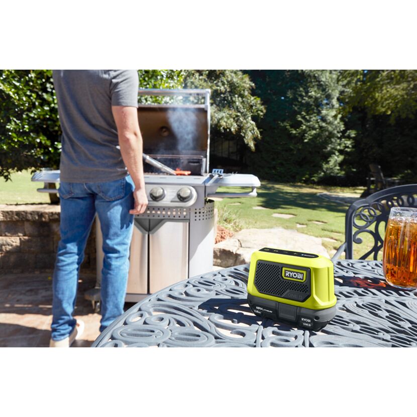 Person grillt im Garten, daneben steht eine Akku Bluetooth Box von Ryobi.