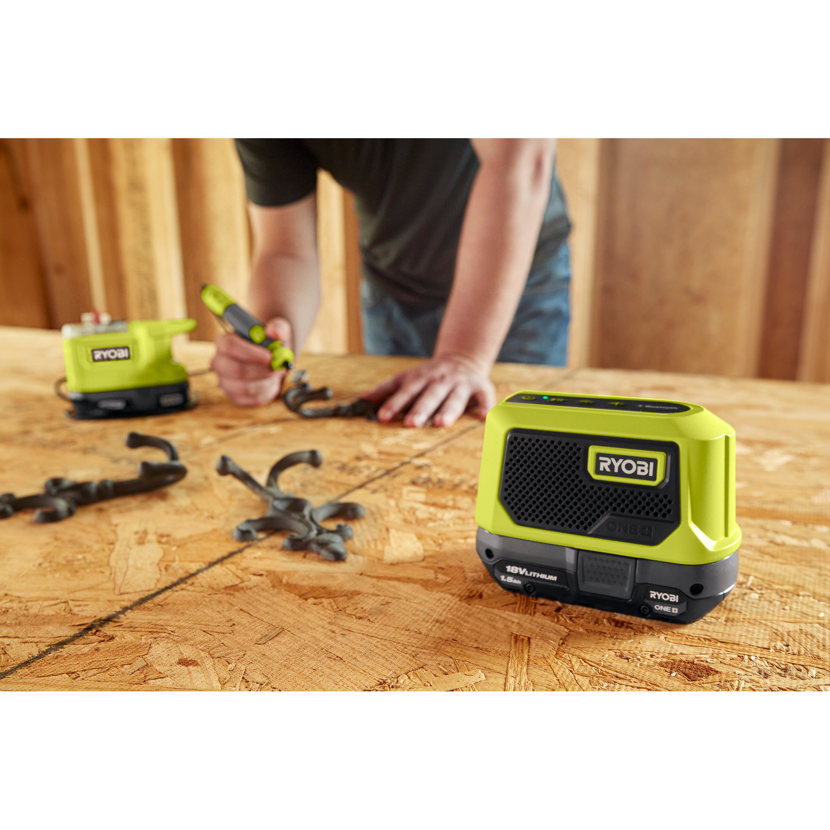 Person bearbeitet mit einem RYOBI Werkzeug ein Werkstück auf dem Tisch. Im Vordergrund die RYOBI Akku Bluetooth Box.