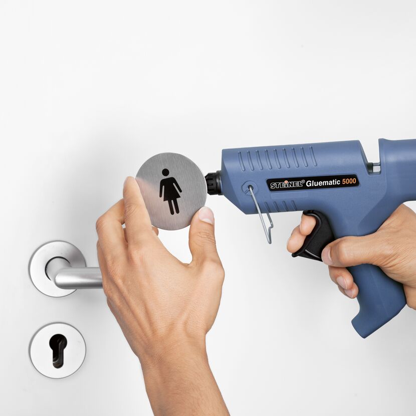 Person bringt mit Heißklebepistole Toilettenschild an