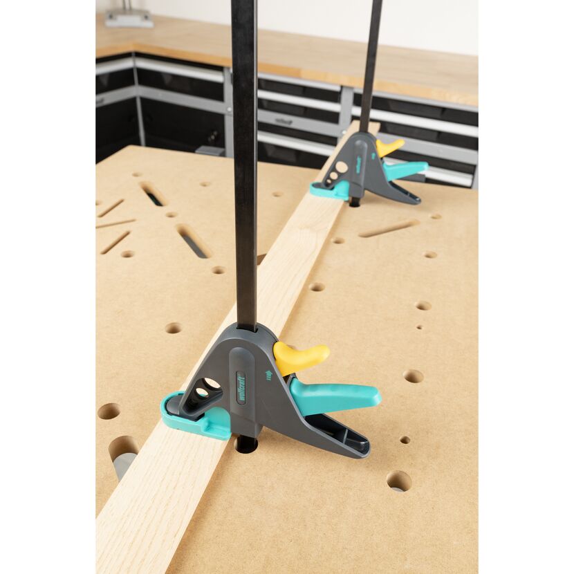 Zwei Einhandzwingen Multi Fix M 150 halten ein Holzstück auf einer Werkbank fest.