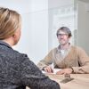 Zwei Personen sitzen an einem Tisch, getrennt durch eine Plexiglasscheibe.