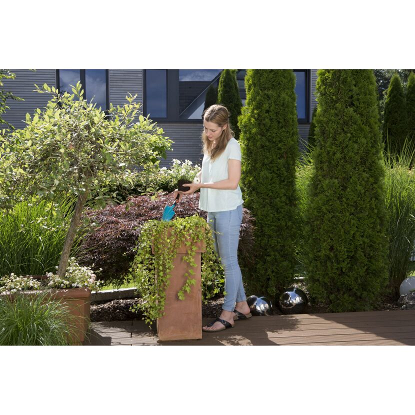 Person pflanzt mit Blumenkelle eine Pflanze in einen Topf.