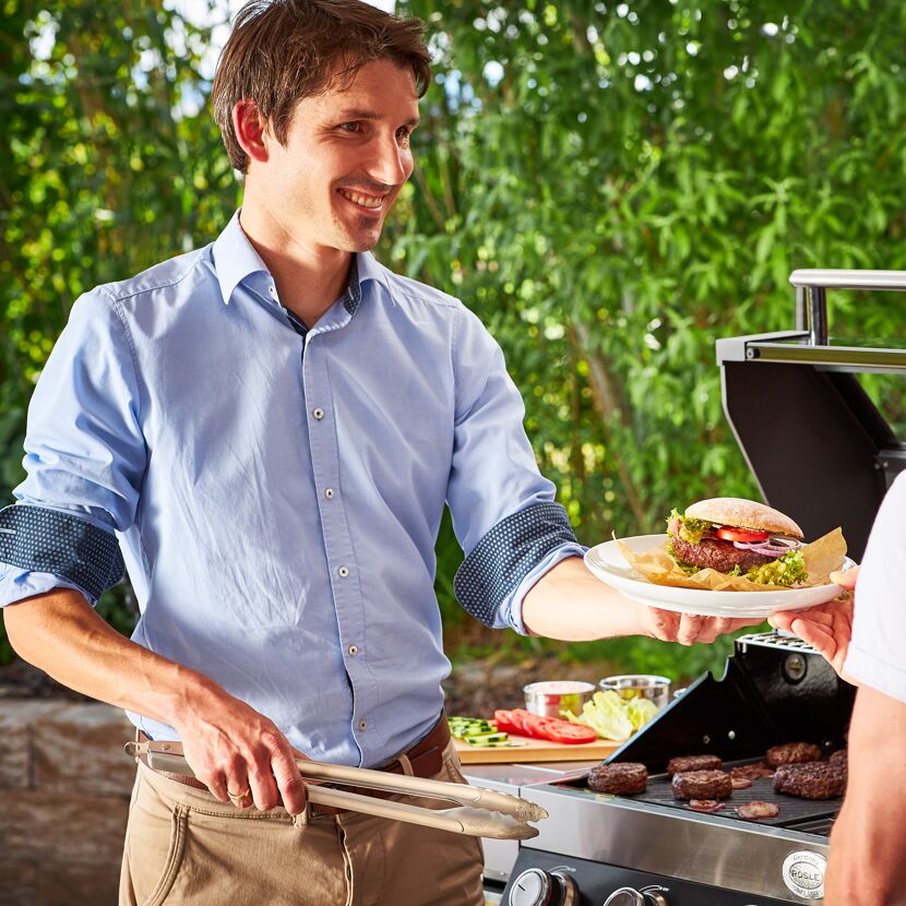 Person grillt Burger und reicht einen Burger mit Grillzange