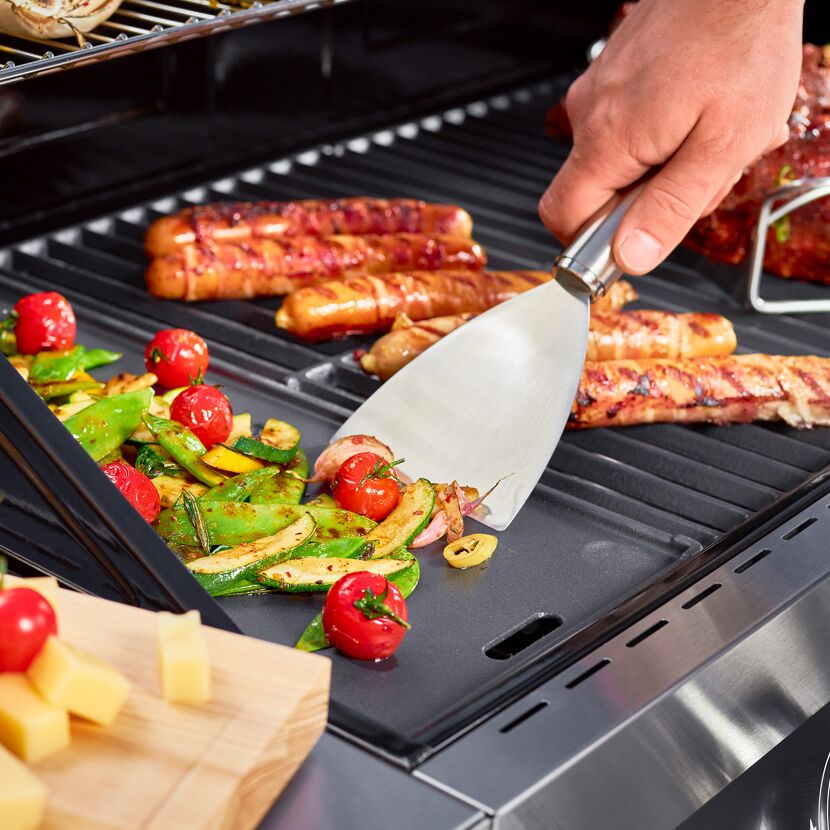 Person grillt Gemüse und Würstchen mit einem Spachtel.