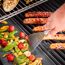 Person grillt Gemüse und Würstchen im Speckmantel mit einem Grillspachtel.