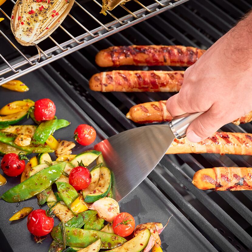 Person grillt Gemüse und Würstchen im Speckmantel mit einem Grillspachtel.