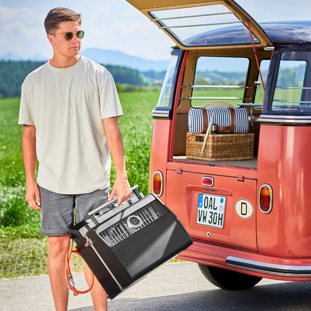 Person trägt einen tragbaren Gasgrill zum VW-Bus.