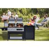 Personen sitzen im Garten und entspannen sich neben einem Gasgrill. Auf dem Grill liegen verschiedene Speisen wie Würstchen, Steaks und Maiskolben.
