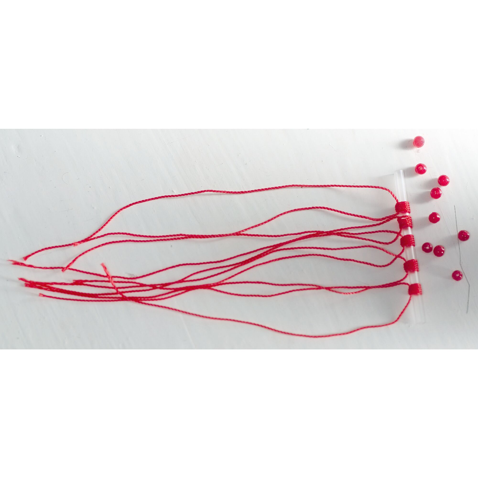 Roter Faden um ein Röhrchen gewickelt. Daneben rote Perlen.