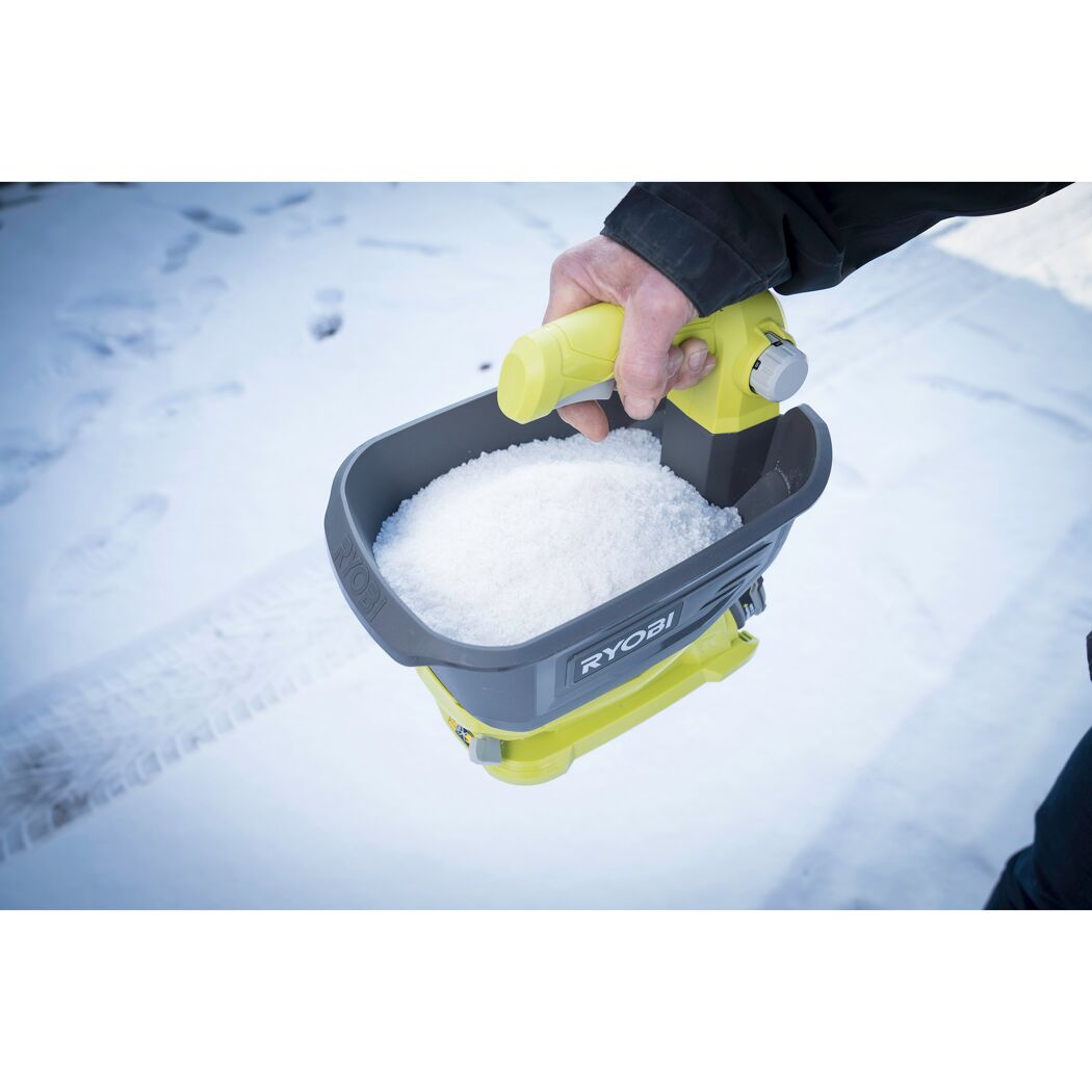 Person hält einen Ryobi Universal-Streuer mit Streugut über verschneitem Boden.