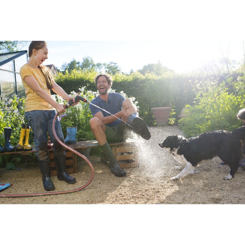 Person spritzt mit einem Gartenschlauch Wasser auf Gummistiefel und Hund