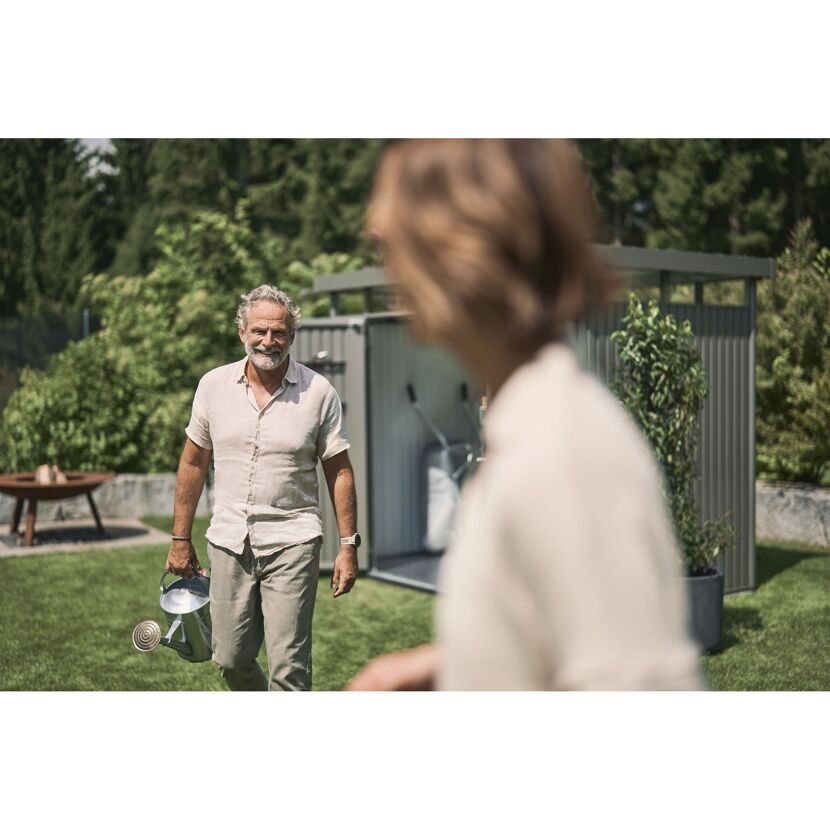 Lächelnde Person trägt Gießkanne im Garten vor Gerätehaus.