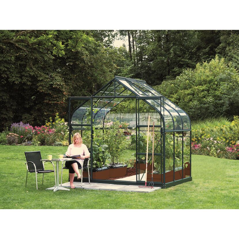 Person sitzt neben einem Gewächshaus mit hochgewachsenen Pflanzen im Garten.