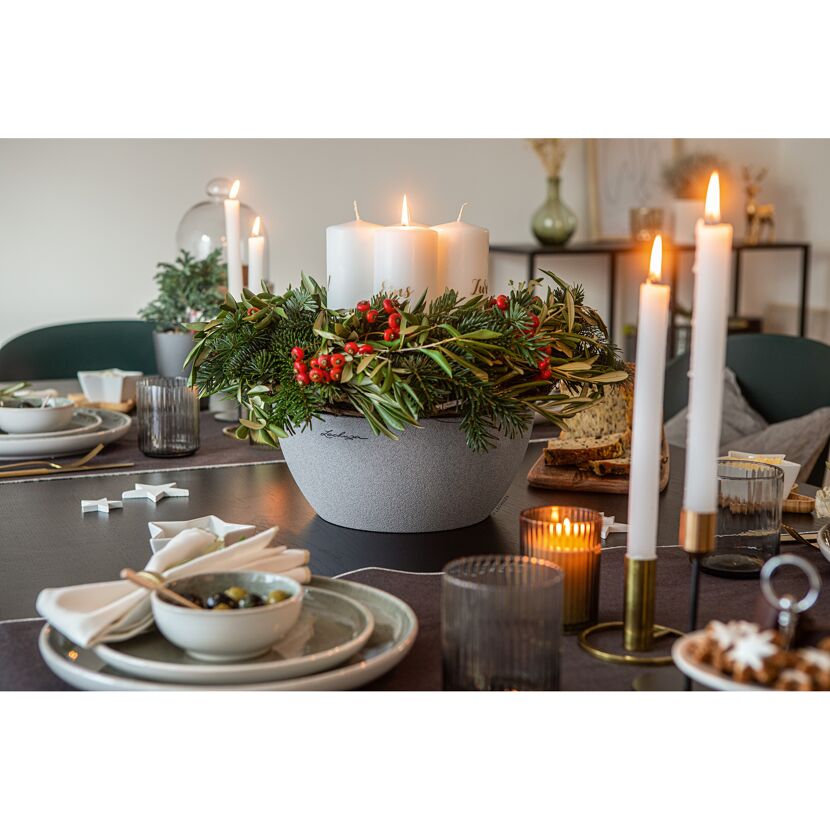 Adventskranz mit Kerzen in grauer Schale auf gedecktem Tisch.