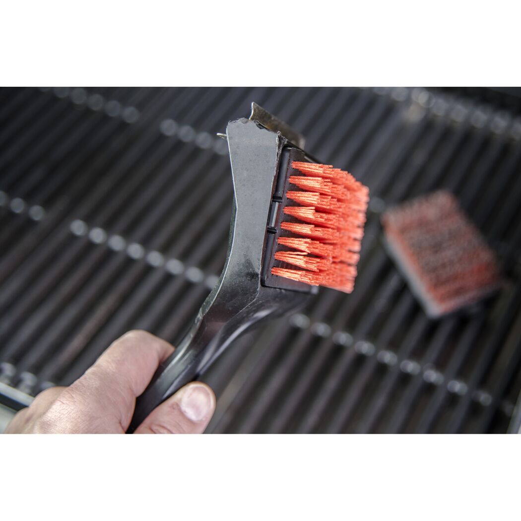Person hält einen Grillreiniger mit orangen Borsten über einem Grillrost.