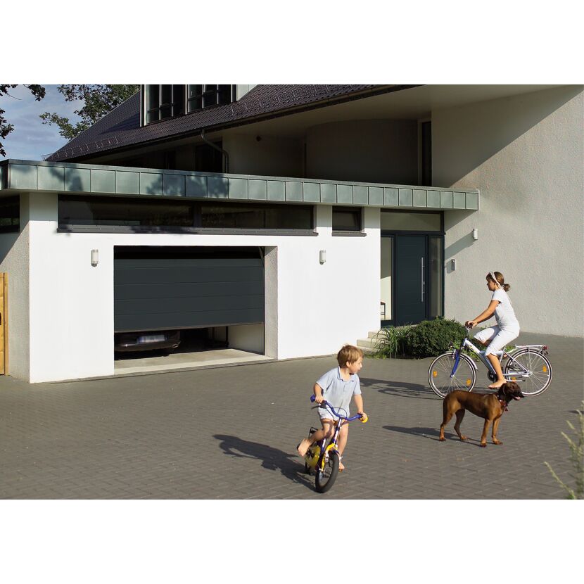 Zwei Kinder und ein Hund fahren Fahrrad vor einem Haus mit geöffnetem Garagentor.