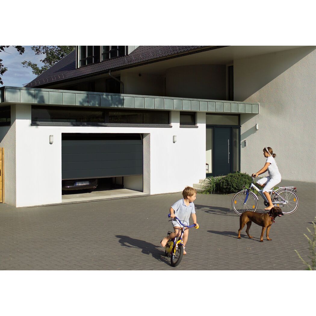 Zwei Kinder und ein Hund fahren Fahrrad vor einem Haus mit Garagentor.