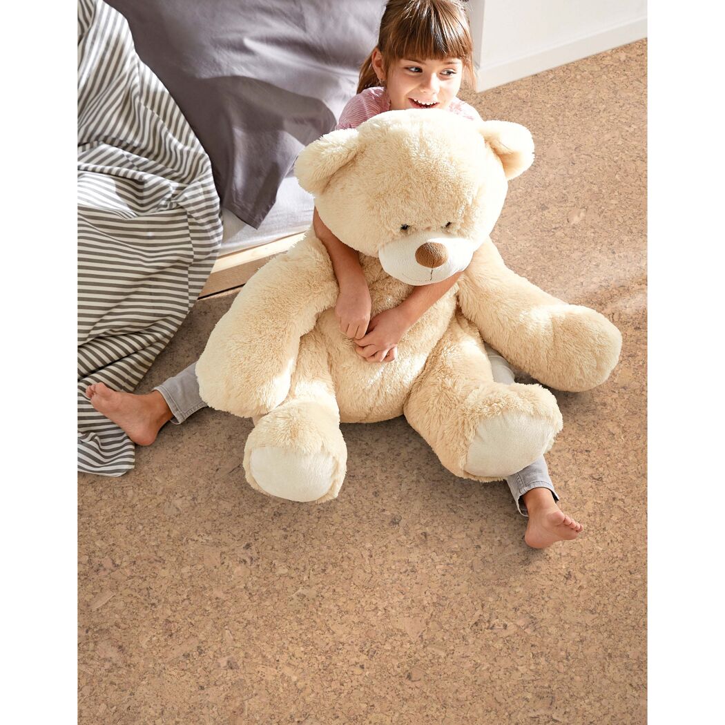 Kind sitzt auf Korkboden und kuschelt mit großem Teddybären.