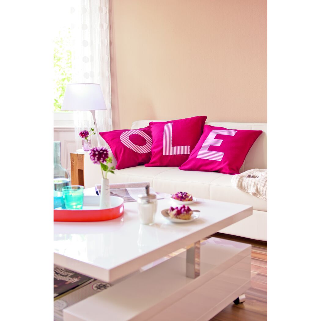 Wohnzimmer mit weißen Sofa und pinken Kissen mit Schriftzug.