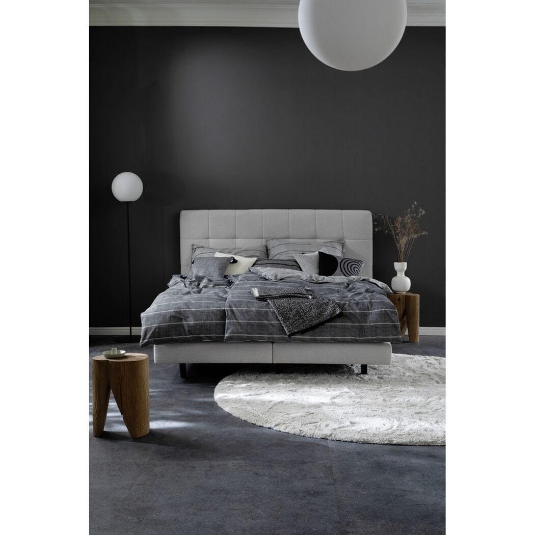 Helles Bett mit grauer Bettwäsche vor schwarzer Wand.