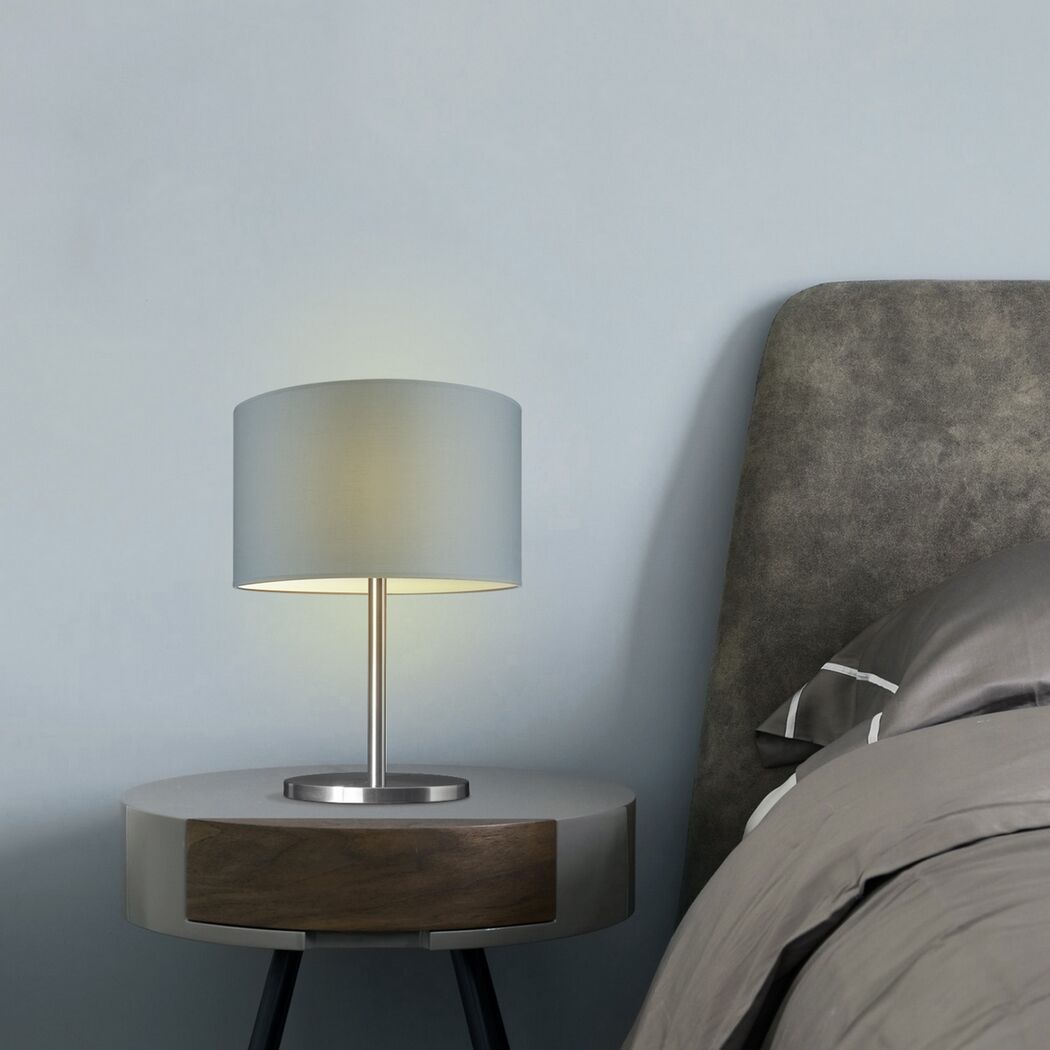 Eine graue Tischlampe auf einem Nachttisch neben einem Bett.